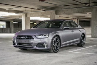 Audi berline en gris nardo LY7C garée dans un parking urbain en béton, vue en trois quarts avant basse révélant la teinte satin caractéristique